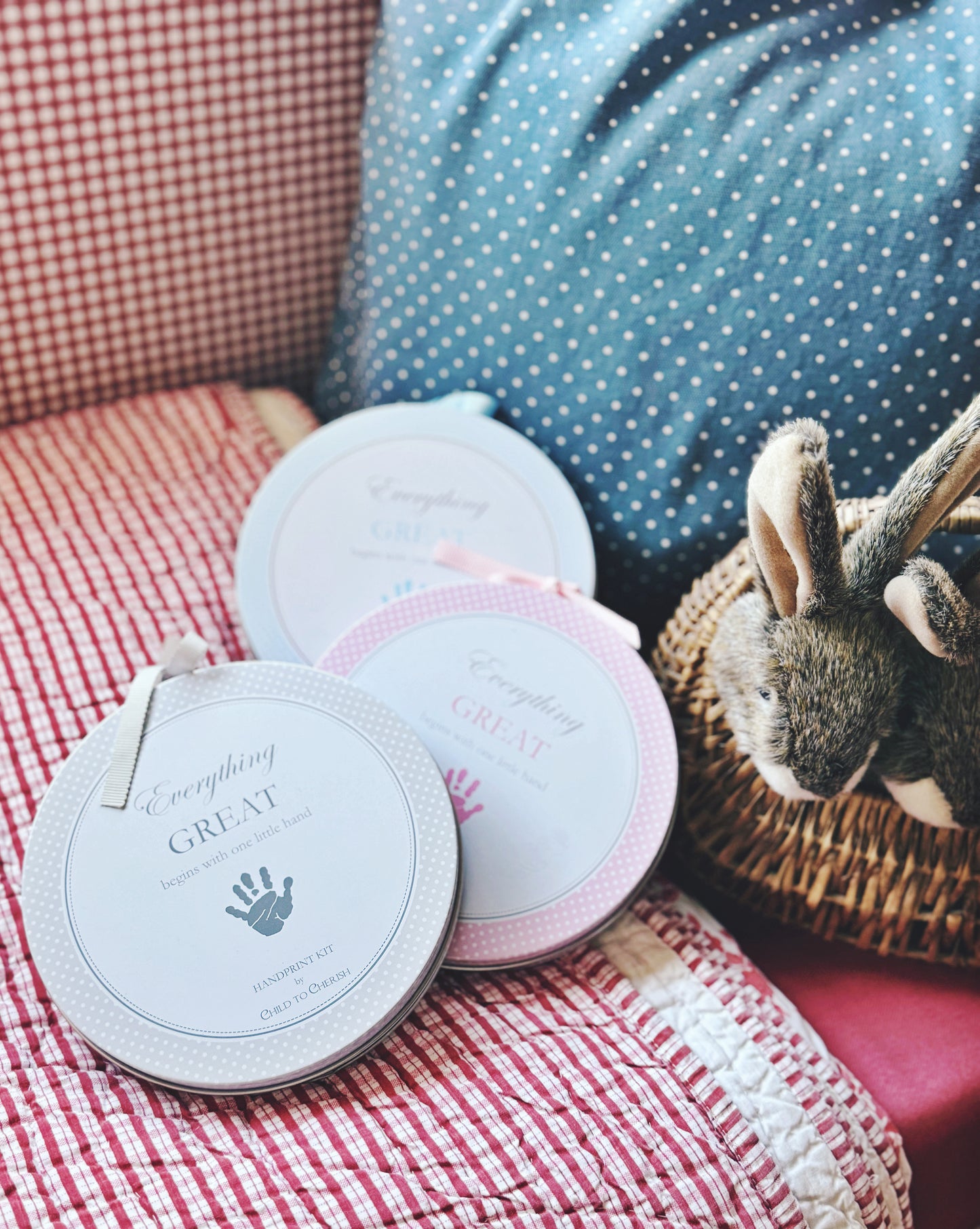 Three round handprint kits in gray, pink, and blue on a cushioned chair with stuffed bunny