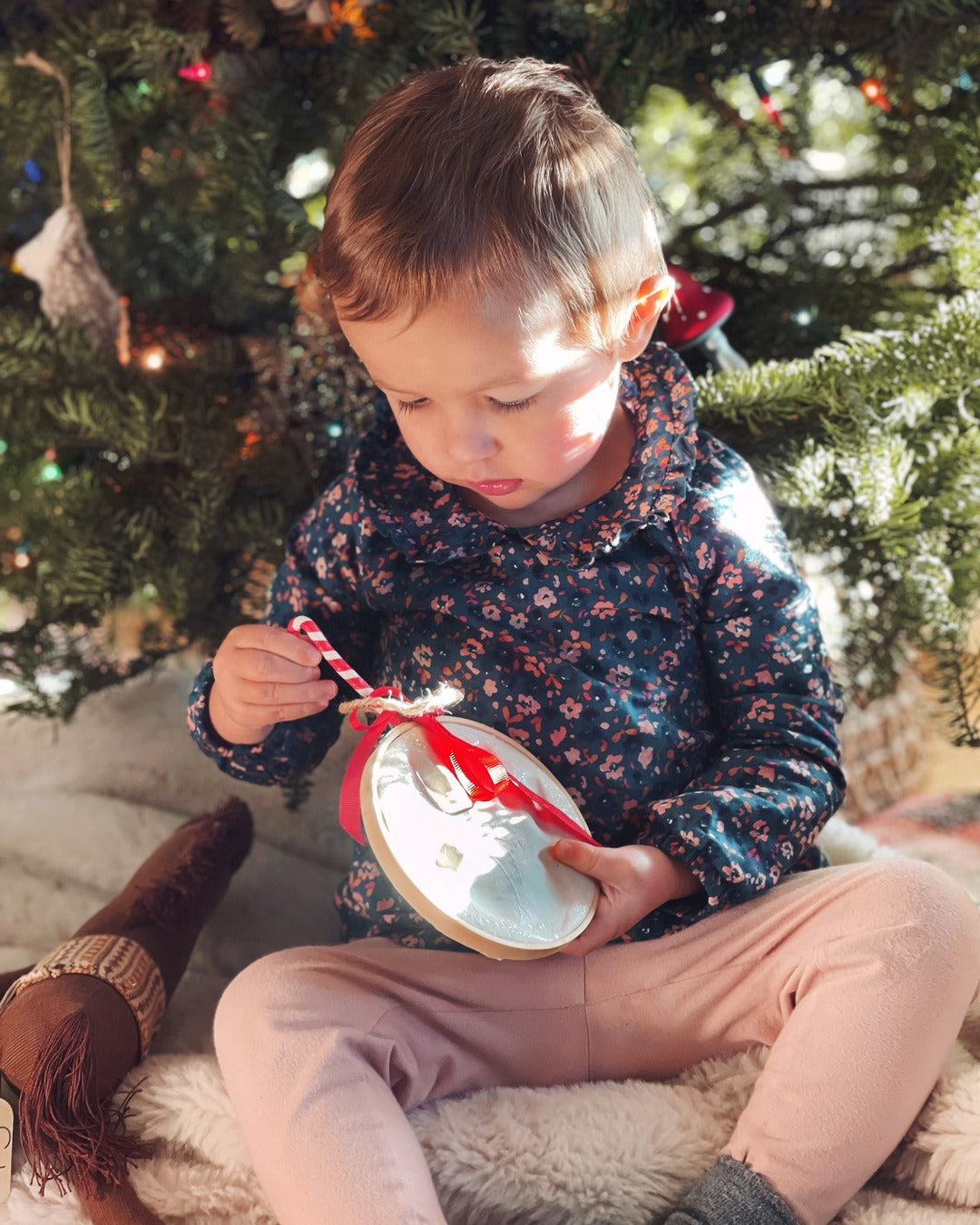 A toddler sitting by the Christmas tree holding a Child to Cherish Claydough handprint ornament