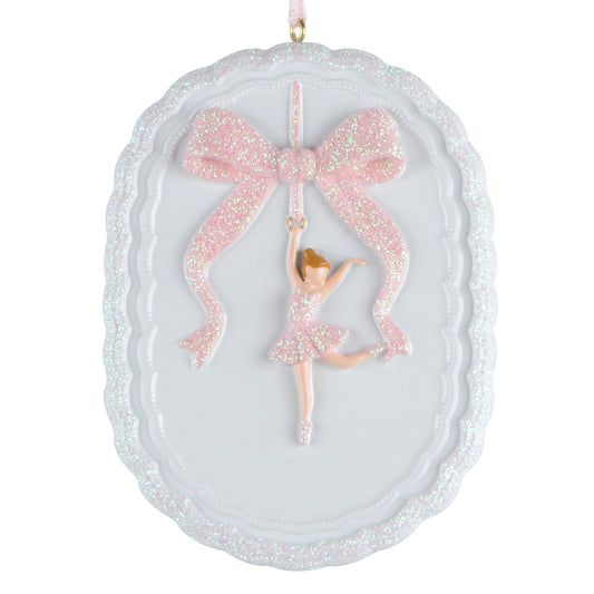 Oval white holiday ornament with scalloped edges featuring a handpainted ballerina in a pink glitter dress hanging from a pink glitter bow