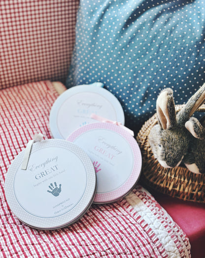 Three round handprint kits in gray, pink, and blue on a cushioned chair with stuffed bunny
