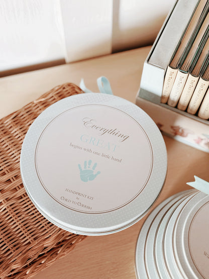 Stack of blue handprint kits in round tins with polka dot borders, placed on woven basket