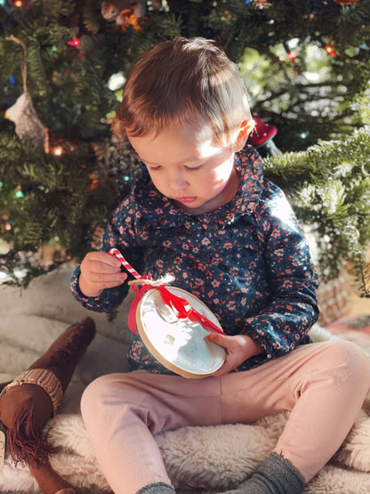 Child holding and looking at the handprint ornament seated by a Christmas tree