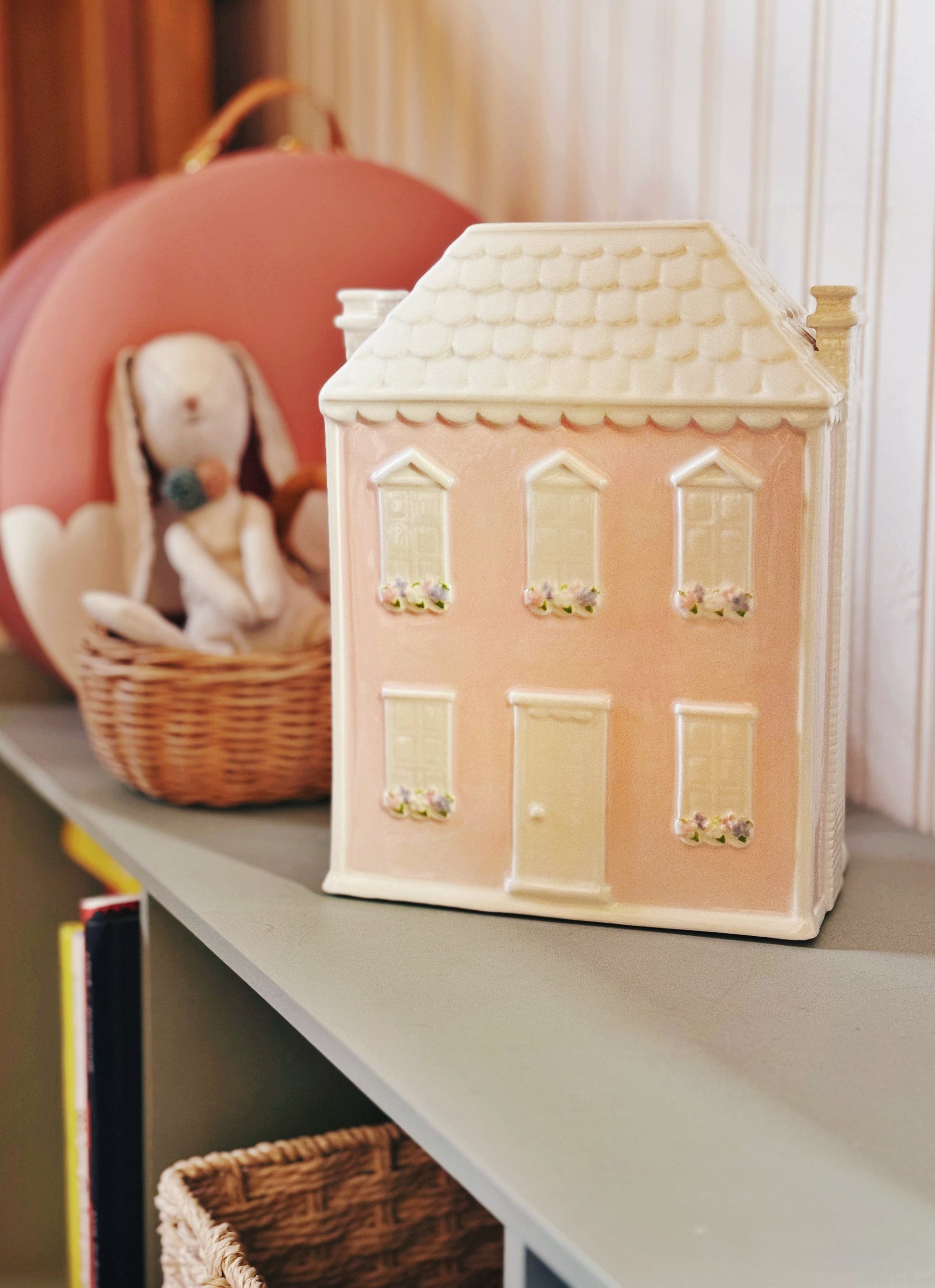 Pink house piggy bank on gray shelf near toy basket and pink suitcase with child placing coins inside