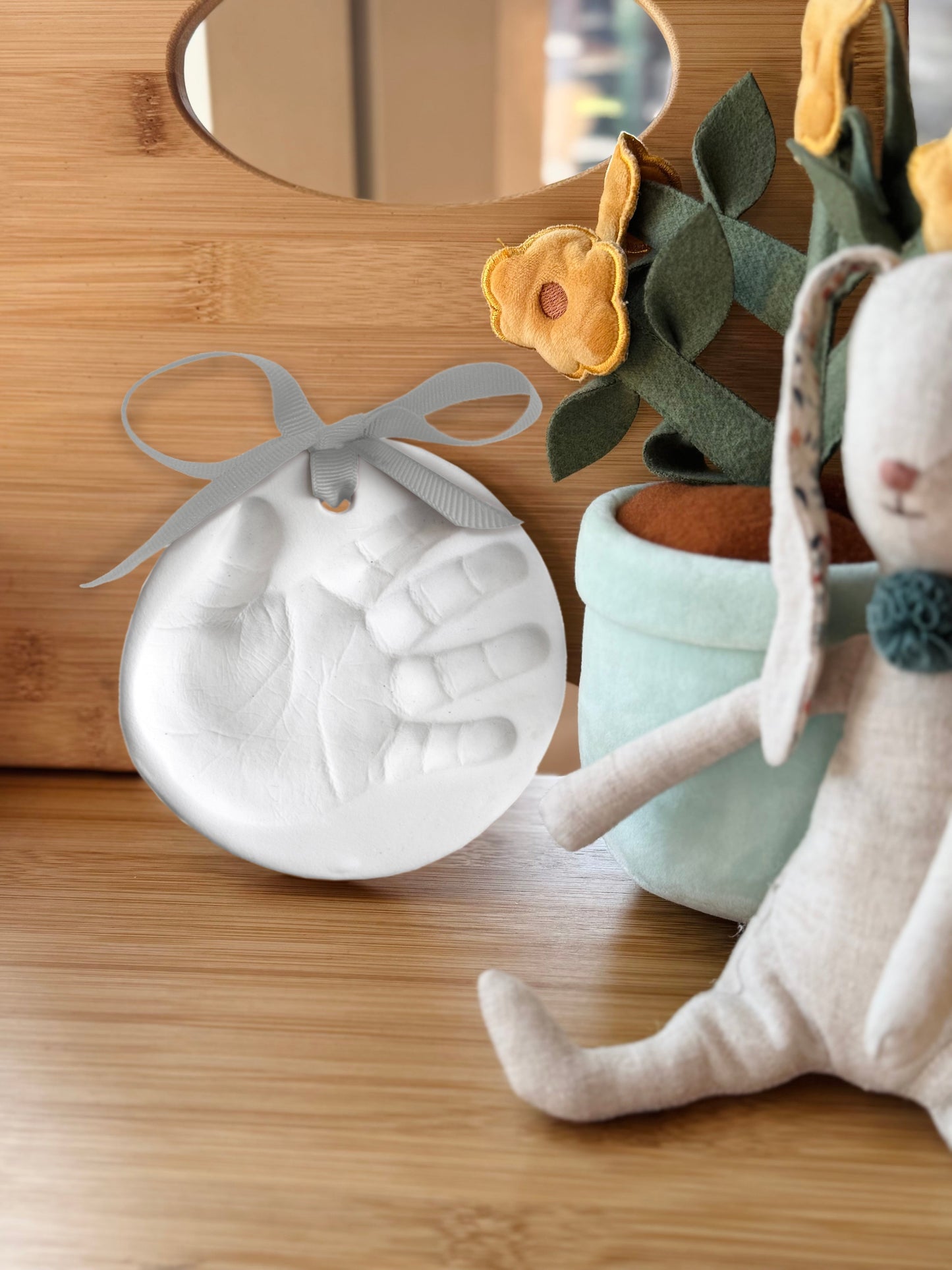Finished clay handprint ornament displayed on wooden shelf next to soft plush toys and felt flowers
