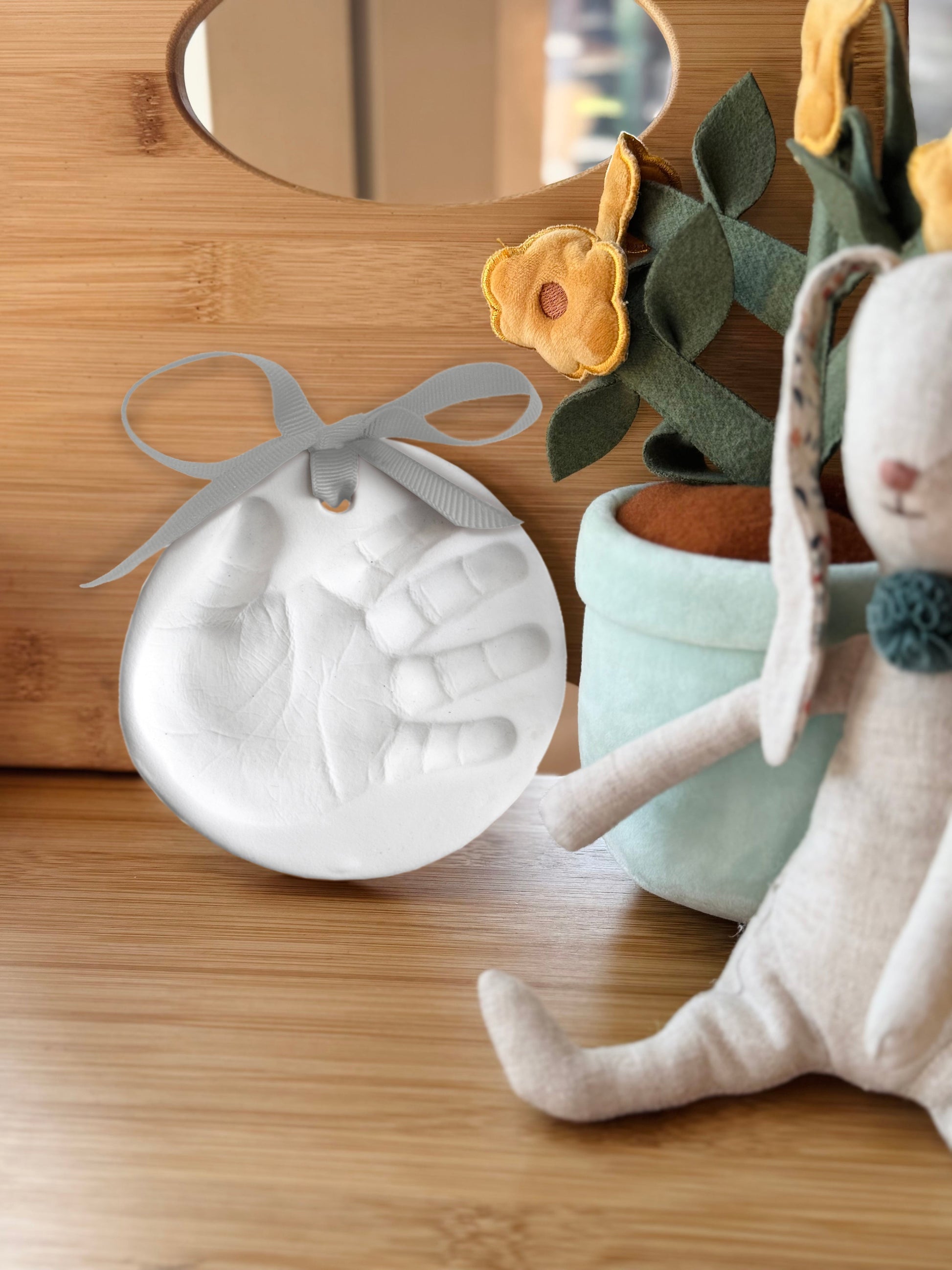 Finished clay handprint ornament displayed on wooden shelf next to soft plush toys and felt flowers