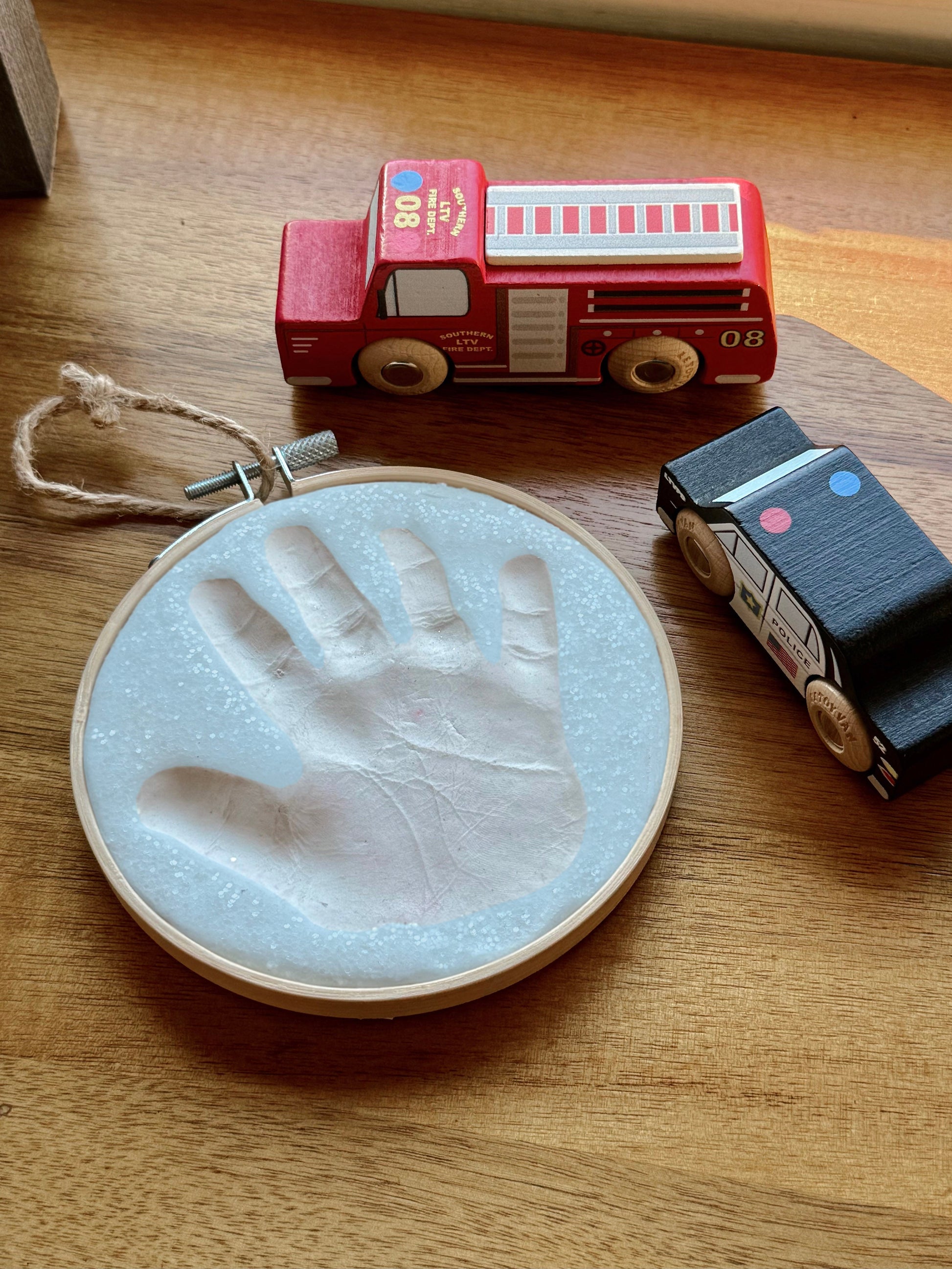 Close-up of blue glitter handprint ornament displayed on wood surface next to wooden toy fire truck and police car