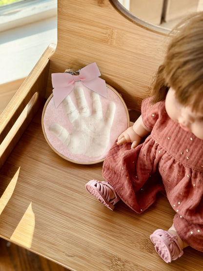 Handprint keepsake displayed on wooden surface next to a doll in a pink dress