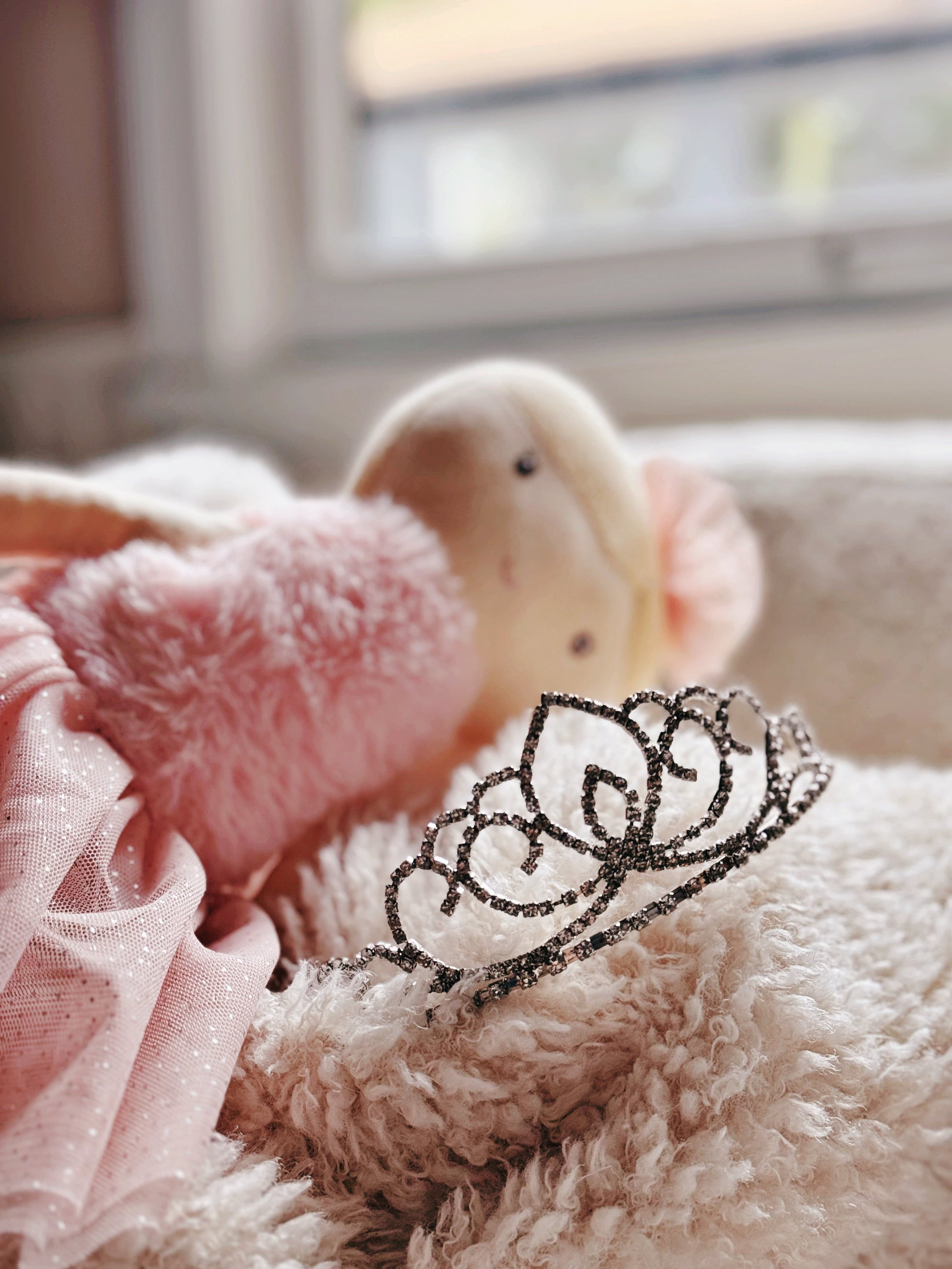 Silver rhinestone tiara lying on a soft white textured blanket with plush doll nearby