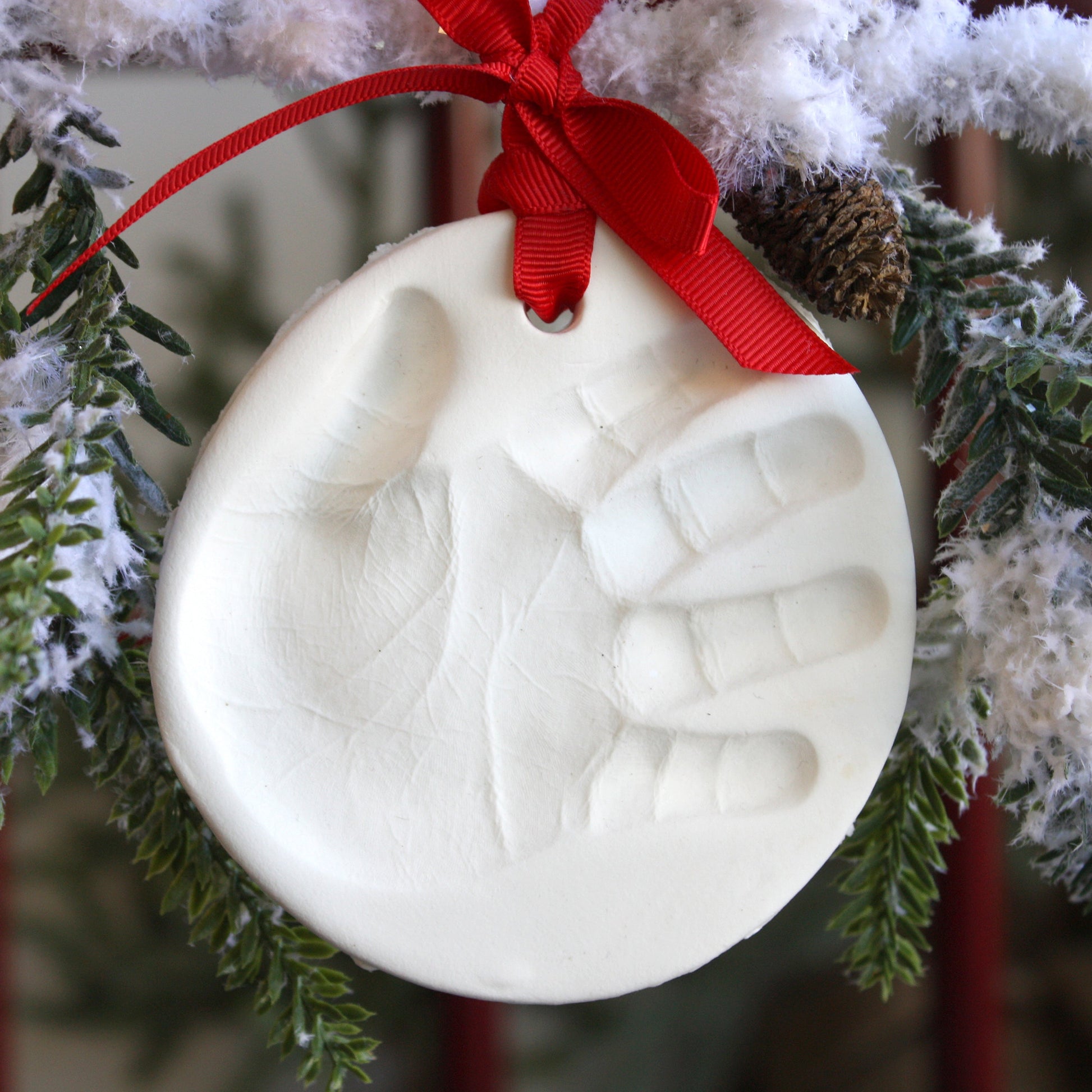 Close-up of completed white clay handprint ornament hanging on snowy Christmas tree branches with red ribbon